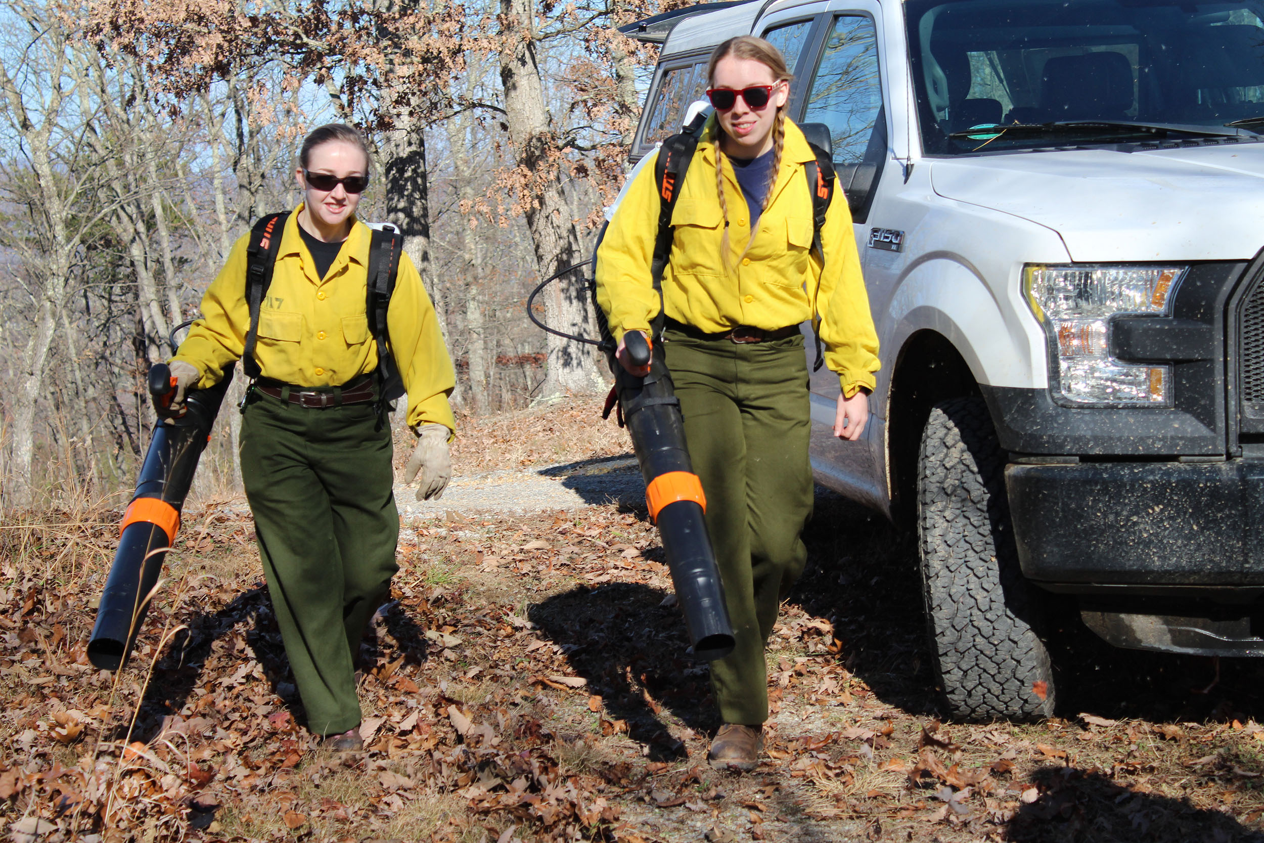 Students Molly Hunt (left) and Emily Newcombe use leaf blowers to clear the fire line of any loose debris in advance of the prescribed burn